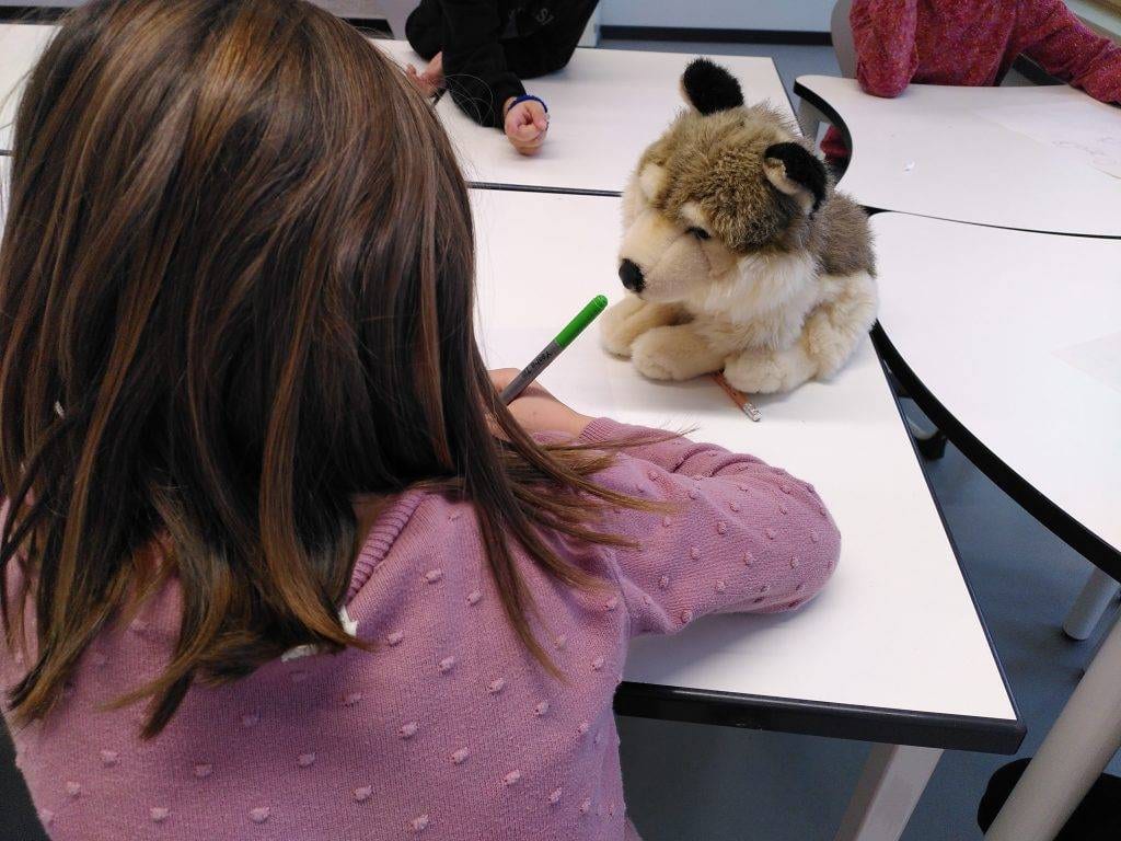 ein kind mit braunen haaren, das einen rosa pullover trägt, schreibt an einem weißen tisch vor einem plüschwolf. andere kinder sitzen in der nähe, teilweise sichtbar, und arbeiten ebenfalls an den tischen.