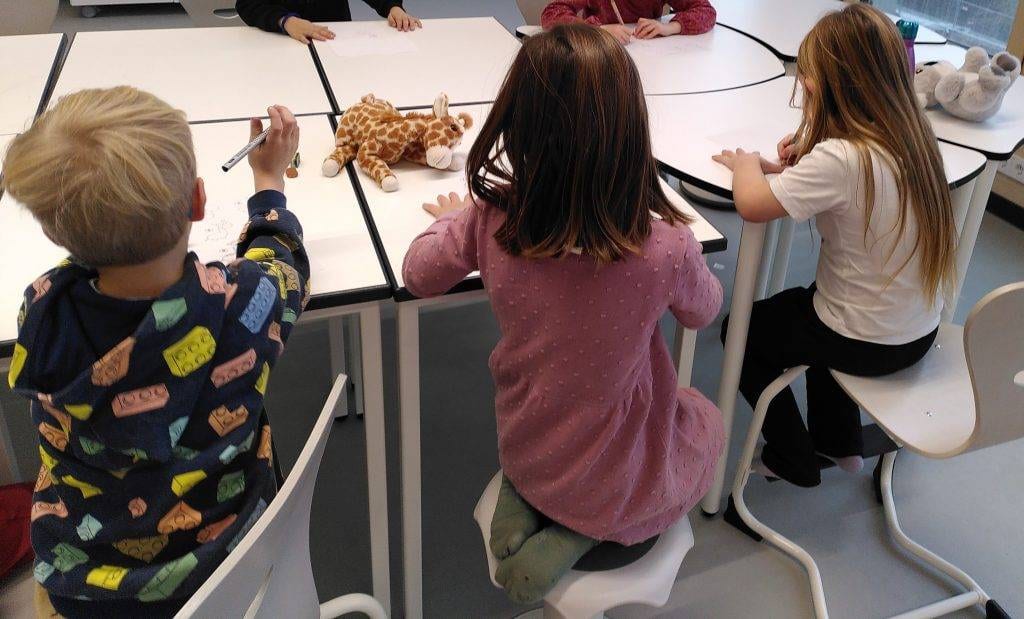 vier kleine kinder sitzen an einem weißen tisch und zeichnen auf papier. zwei plüschtiere, eine giraffe und ein elefant, liegen auf dem tisch. die kinder werden von hinten betrachtet und scheinen auf ihre tätigkeit konzentriert zu sein.