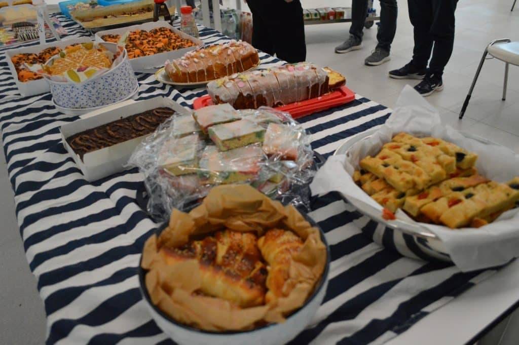 ein mit einem gestreiften tuch bedeckter tisch ist mit verschiedenen backwaren, darunter kuchen, focaccia, gebäck und sandwiches, belegt. mehrere personen stehen um den tisch herum, ihre beine sind im hintergrund zu sehen.