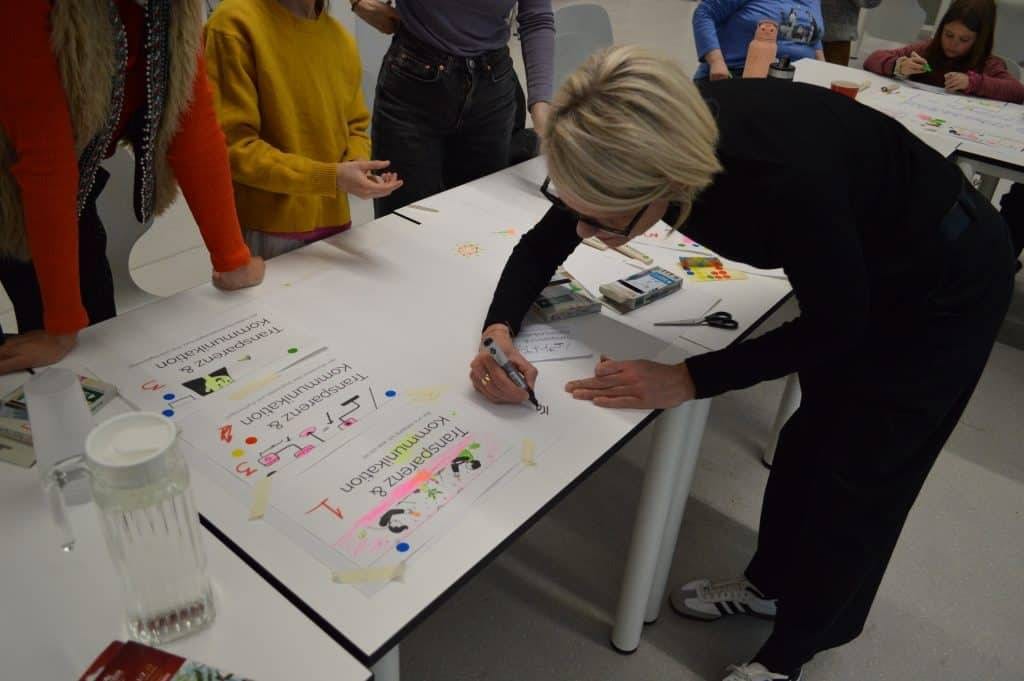 eine frau beugt sich über einen tisch und schreibt auf ein großes blatt papier mit bunten zeichnungen und texten. mehrere personen stehen oder sitzen in der nähe und beschäftigen sich ebenfalls mit kreativen tätigkeiten.
