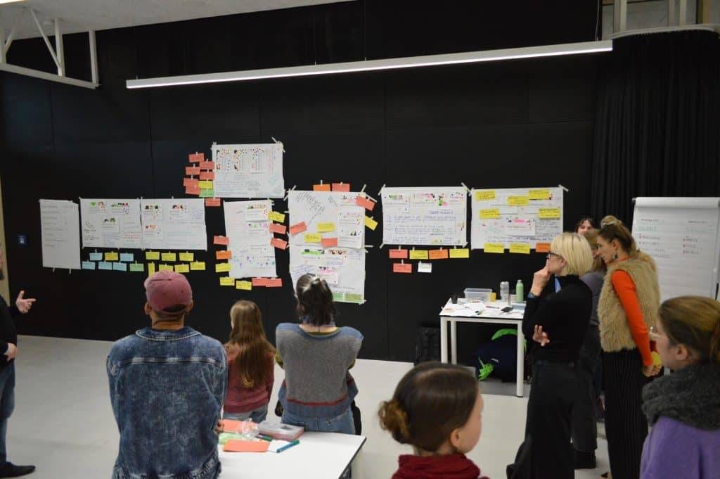 eine gruppe von personen steht in einem raum vor einer schwarzen wand mit großen papieren, tabellen und bunten haftnotizen, die in reihen angeordnet sind, als ob sie an einem workshop oder einer gemeinsamen brainstorming sitzung teilnehmen würden.
