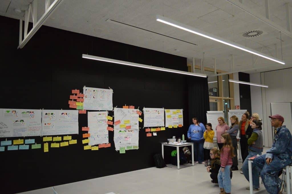 eine gruppe von menschen, darunter kinder und erwachsene, steht und sitzt vor einer schwarzen wand, die mit bunten postern und haftnotizen in einem modernen, gut beleuchteten raum bedeckt ist.