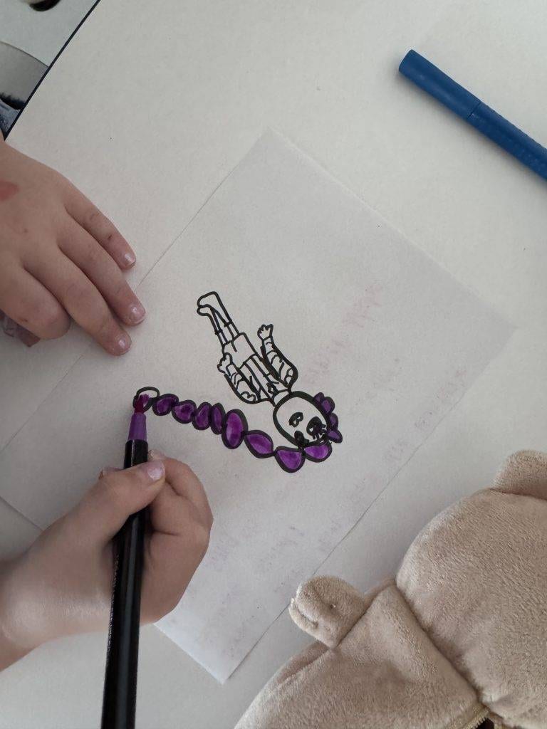 ein kind malt die zeichnung einer figur auf einem weißen blatt papier mit einem lila marker aus. ein weiterer blauer marker und ein plüschbär liegen in der nähe auf dem weißen tisch.