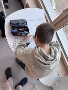 ein kind sitzt an einem weißen schreibtisch und zeichnet mit buntstiften. auf dem tisch liegen ein offenes federmäppchen und papiere mit zeichnungen. das kind trägt einen hellgrauen kapuzenpullover und arbeitet in der nähe eines großen fensters, in dessen nähe eine weitere person steht.