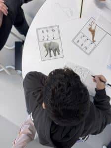 ein kind sitzt an einem runden weißen tisch und malt ein tier auf papier. auf dem tisch liegen tierkarten mit musiknoten und abbildungen eines elefanten, eines dinosauriers und einer giraffe. andere kinder sind teilweise sichtbar.