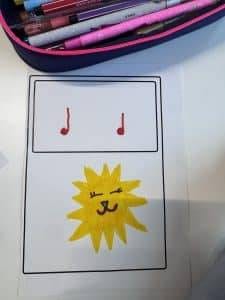 ein blatt papier zeigt oben in einem kasten zwei rote musiknoten und unten in einem kasten die zeichnung einer lächelnden, zwinkernden gelben sonne mit einem niedlichen gesicht. am oberen rand des bildes sind rosa stifte und ein federmäppchen zu sehen.