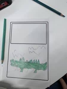 die zeichnung eines kindes auf einem arbeitsblatt zeigt unten einen grünen, stacheligen dinosaurier oder drachen und darüber berge und formen. auf dem weißen tisch sind zwei grüne bleistifte und ein teil einer federmappe zu sehen.