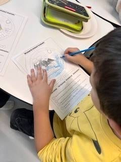 ein kind in einem gelben pikachu shirt malt mit einem blauen farbstift ein arbeitsblatt mit dem titel 