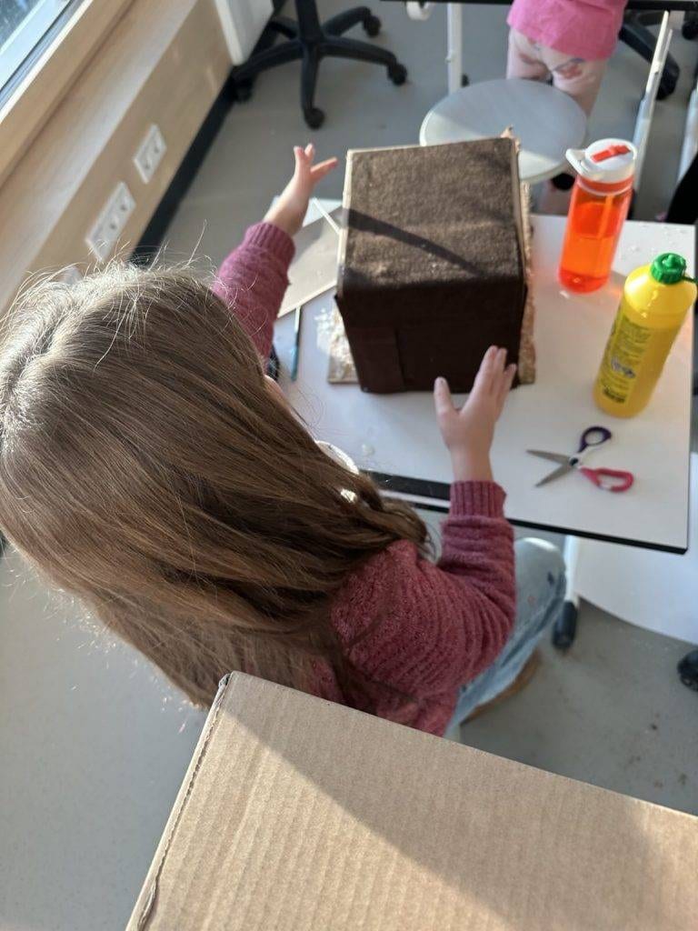 ein kind mit langen braunen haaren und einem rosa pullover sitzt an einem tisch und arbeitet an einem braunen karton. auf dem tisch liegen eine schere, eine wasserflasche und eine gelbe flasche. das sonnenlicht fällt durch das fenster.