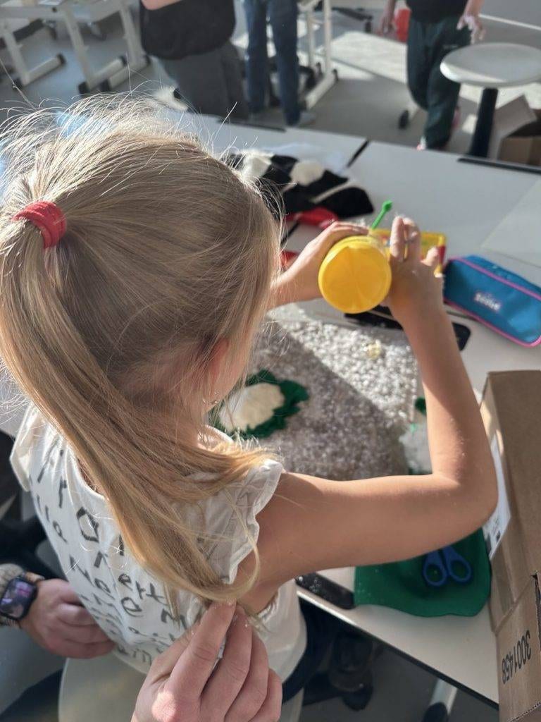ein junges mädchen mit blondem haar in einem pferdeschwanz arbeitet an einem bastelprojekt an einem tisch und schüttet gelbes pulver auf eine strukturierte oberfläche, umgeben von bastelmaterial und anderen schülern in einem hell erleuchteten klassenzimmer.