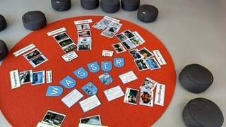 ein runder orangefarbener teppich, auf dem das wort wasser in blauen karten geschrieben steht, umgeben von verschiedenen fotos und textkarten. graue kissen sind im kreis um den teppich angeordnet.