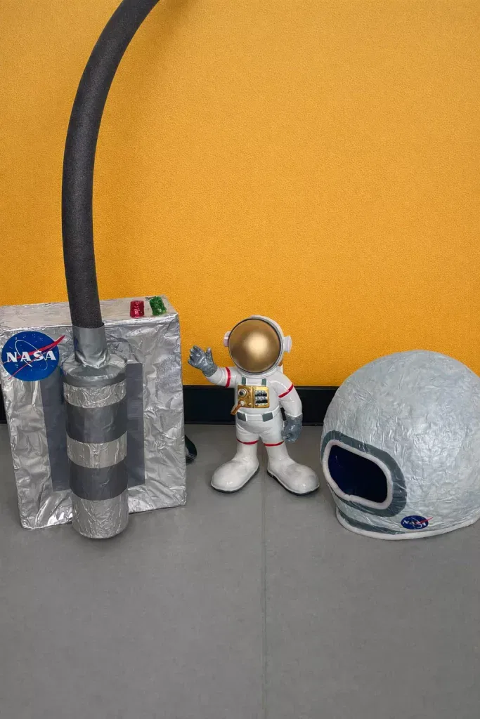 eine kleine astronautenfigur steht zwischen einer selbstgemachten silbernen schachtel mit nasa logos und knöpfen und einem großen grauen helm, alles vor einer gelben wand auf einem grauen boden.