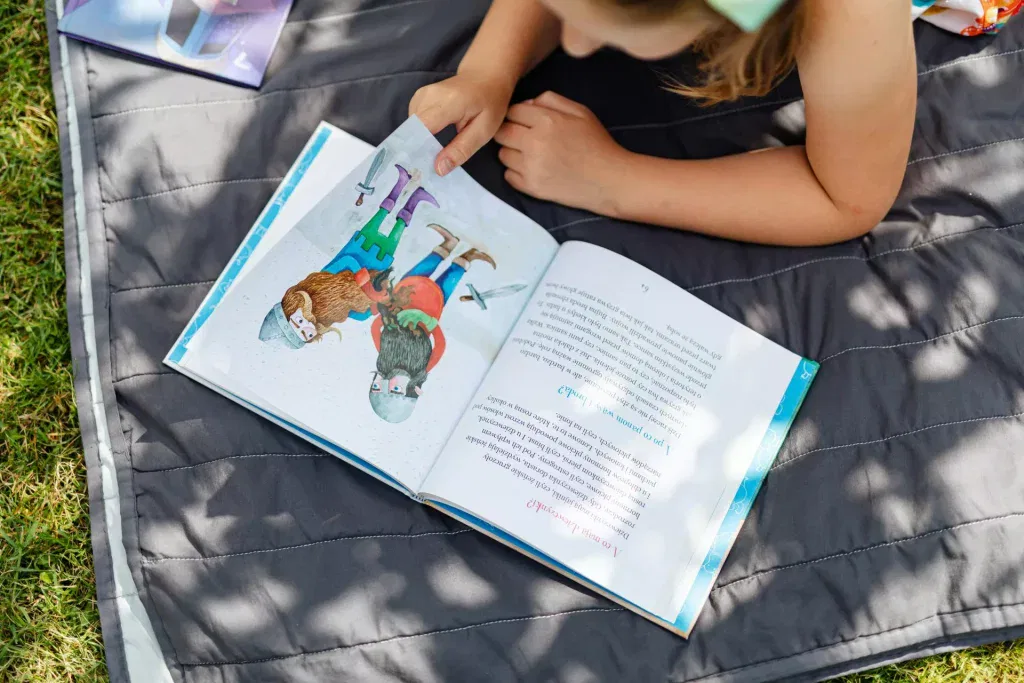 ein kind liegt auf einer grauen decke im freien und liest ein illustriertes buch, das zwei mädchen mit tieren zeigt. das sonnenlicht fällt durch die Äste der bäume und wirft schatten auf die seite und die decke.
