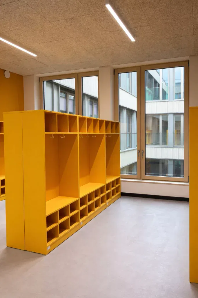 leuchtend gelbe fächerschränke mit regalen und haken sind in einem modernen klassenzimmer oder kindertagesstättenraum mit großen fenstern und viel natürlichem licht in reihen angeordnet.