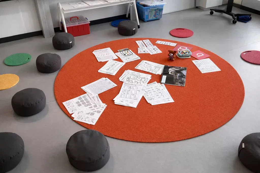 ein runder orangefarbener teppich, auf dem arbeitsblätter, ein buch und kleine spielsachen verstreut liegen. graue bodenkissen umgeben den teppich, und im hintergrund des klassenzimmers befinden sich bunte matten und aufbewahrungsboxen.