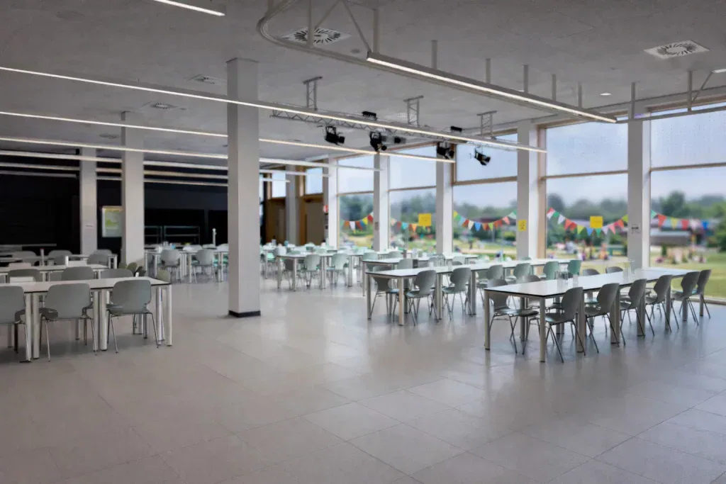 eine geräumige, moderne cafeteria mit reihen leerer weißer tische und stühle, großen fenstern, die natürliches licht hereinlassen, und bunten wimpeln, die draußen auf einer wiese zu sehen sind.