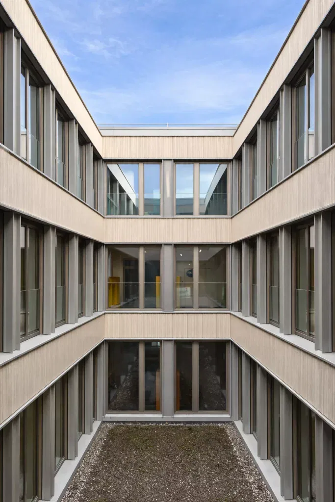 ein modernes, dreistöckiges gebäude mit großen fenstern und einem zentralen, offenen, mit kies gefüllten innenhof, der zum blauen himmel hinaufragt. die fassade ist beige mit vertikalen metallakzenten.