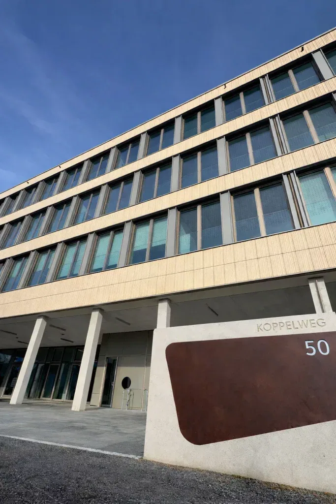 ein modernes beige graues gebäude mit vielen fenstern, von einem niedrigen winkel aus gesehen. davor steht ein betonschild mit der aufschrift koppelweg 50 und einem großen braunen akzent. der himmel über dem gebäude ist klar und blau.