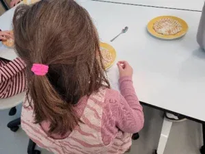 ein junges mädchen mit braunen haaren, die zu einem rosa haargummi gebunden sind, sitzt an einem weißen tisch mit einem gelben teller, auf dem eine waffel liegt. ein anderes kind ist teilweise neben ihr zu sehen, ebenfalls mit einer waffel auf einem gelben teller.
