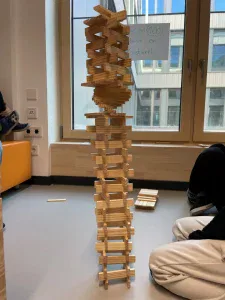ein hoher, verschlungener turm aus holzbrettern steht in einem innenraum in der nähe eines fensters, umgeben von menschen, die auf dem boden sitzen. der turm weist gestaffelte und ineinandergreifende schichten auf, mit einer runden struktur nahe der spitze.