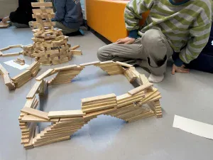 ein kind in einem grün gestreiften pullover sitzt auf dem boden in der nähe von holzklötzchenstrukturen, darunter ein rundbogen und ein turm mit der aufschrift kapla. im hintergrund sind weitere kinder und klötze zu sehen.