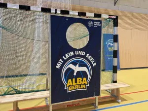 ein schild mit dem logo der basketballmannschaft alba berlin und der aufschrift "mit leib und seele" steht vor einem tor in einer sporthalle. im hintergrund sind sportnetze, eine bank und eine holzwand zu sehen.