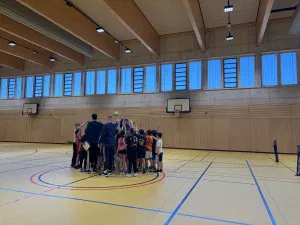 eine gruppe von kindern und zwei erwachsene stehen mit erhobenen armen in einem kreis in einer turnhalle mit hohen fenstern, holzwänden und basketballkörben.